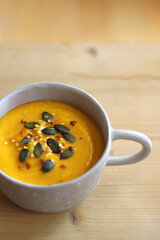 Bowl of creamy pumpkin and potato soup, with pumpkin seeds and chilli flakes toppings. Selective focus, wooden background.