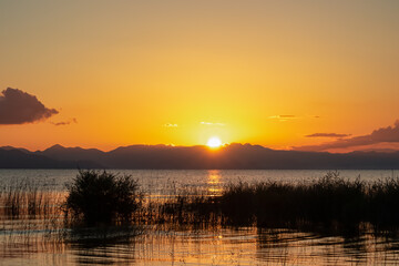 Obraz premium Breathtaking sunset over calm Skadar lake, Albania. Sun is setting behind Dinaric Alps mountains in Montenegro. Water reflects vibrant colors of sky creating shimmering path across the surface
