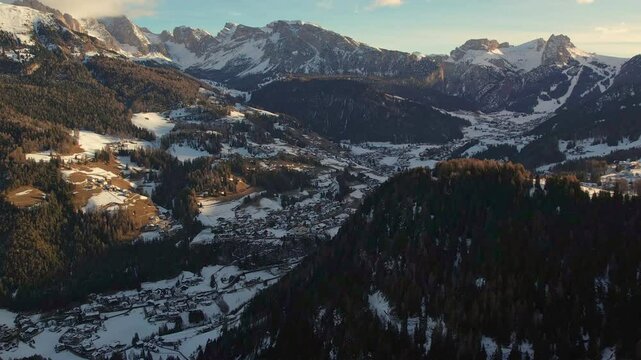 A beautiful aerial view of Wolkenstein an alpine village nestled among snow-dusted dolomite mountains and forests. The setting sun casts a warm glow on the winter landscape.
