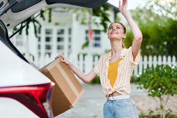 Girl unloading boxes from car trunk in sunny outdoor setting with greenery and white picket fence, showcasing joy and energy in a casual, cheerful atmosphere