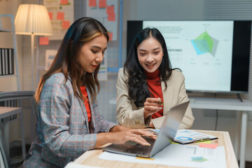 Two businesswomen are collaborating on a laptop over a desk in an office at night, analyzing data and working on a project