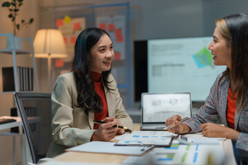 Two businesswomen smiling and talking about work while sitting at a desk in the office at night, analyzing financial data and planning a new project