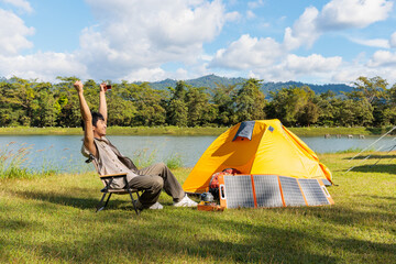 An Asian man feeling fresh and happy after sitting and drinking coffee near camping tent beside the lake and mountain in the sunshine morning with backpack and mobile solar cell