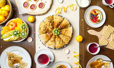 Festive breakfast spread with crepes, fruit tart, and tea. Delicious breakfast with fresh fruit, pancakes, cookies, and tea. Colorful table setting with homemade pancakes, tart, and fruit.