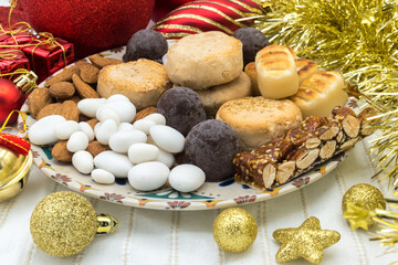 Plate with traditional Christmas sweets in Spain: turr&oacute;n, marzipan, peladillas (sugared almonds), mantecados, polvorones, chocolate balls and almonds. Red and golden decoration on a white tablecloth.