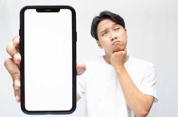 an overthinker young asian man thinking deeply while showing his big screen phone after receiving bad news. mock up of Asian man showing big phone screen while questioning and wondering.