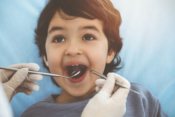 Cute arab boy sitting at dental chair with open mouth during oral checking up with doctor. Visiting sunny dentist office