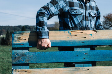 Hand gripping rustic blue pallet, outdoors with plaid shirt and countryside view. Close-up of hand...
