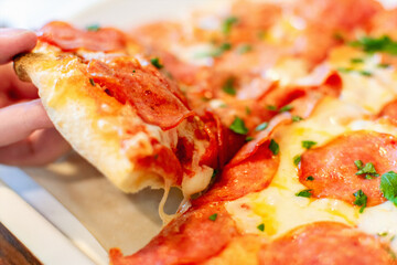 Fresh hot delicious pepperoni pizza with cheese, tomatoes and basil leaves in a pizzeria close-up. Hand taking slice of baked dough and vegetables are traditional Italian cuisine.