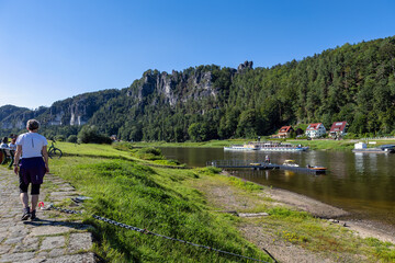 A Woman in Hiking Clothes Strolls Along a Brick Path Beside the River, Where a Steamboat Floats and...