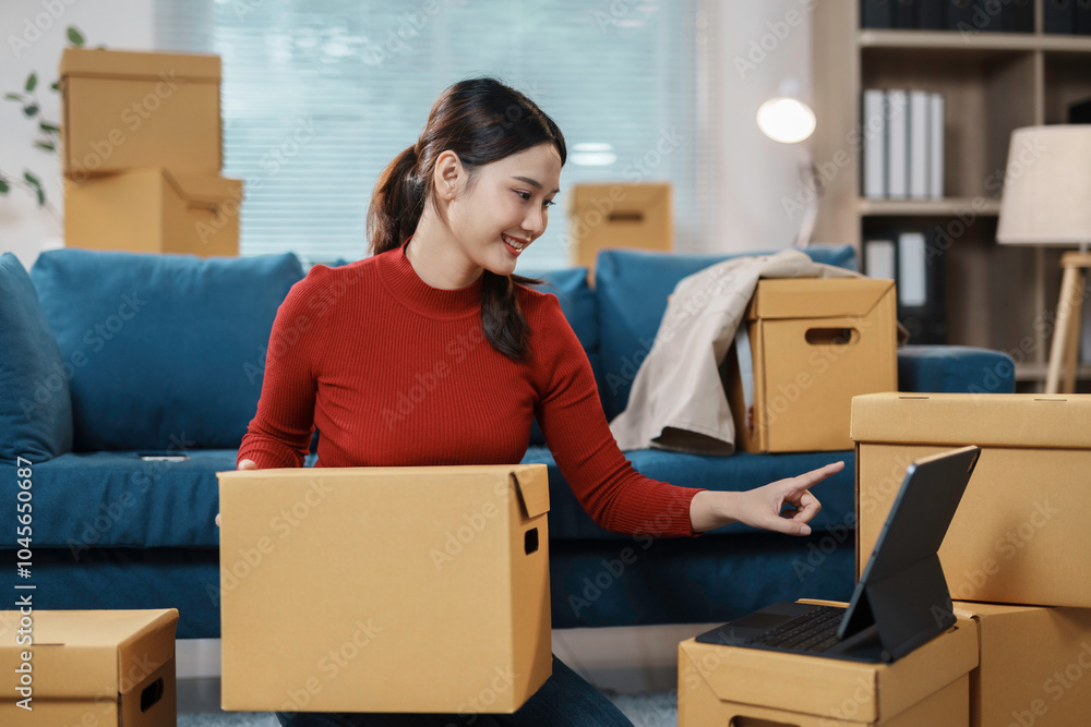 Wall mural young woman is holding a cardboard box and having a video call on a tablet in her living room, surro