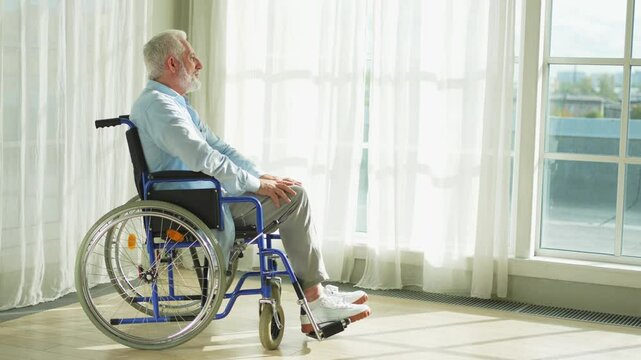Sad senior man sitting in wheelchair looking out window in retirement home hospital. Old paralyzed man in chair for people with disability feel depressed. Lonely elderly sick man sitting on wheelchair