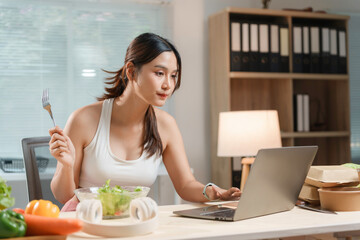 Young asian woman is enjoying a healthy salad while working on her laptop in her home office