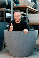 Caucasian man smiles, sitting inside a large plant pot in an outdoor garden store during daylight. A man gives a thumbs-up, ok while sitting comfortably inside a large flowerpot at an outdoor store