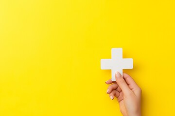 Fototapeta premium Minimalist hand holding a white plus symbol against a bright yellow background, perfect for conceptual designs, healthcare, positivity, medical, and teamwork inspired themes and marketing materials. 