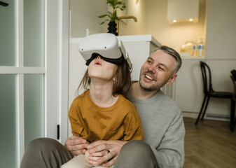 A father watches with amusement as his daughter experiences virtual reality for the first time. She...