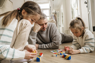 A father lies on the floor, watching his two daughters play with colorful wooden blocks. The girls...