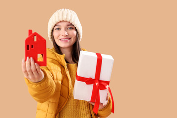 Young woman in winter clothes with Christmas gift and candle holder on beige background