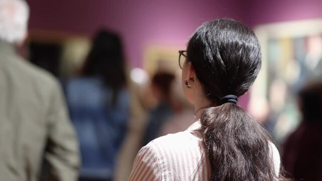 Back view of pretty Caucasian woman wear eyeglasses and listen to guide. Excursion to museum or an art gallery. Museum day and cultural education