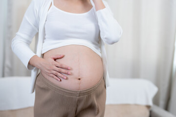 Asian pregnant woman adjusting headphones on baby bump in softly lit room. Calm expression showing connection with unborn child, enjoying soothing music during late pregnancy.