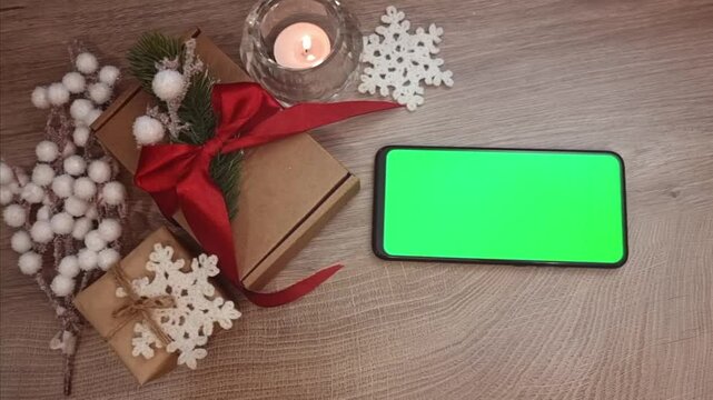 Green screen smartphone lying on wooden table horizontally next to burning candle, gifts fnd DIY Christmas decorations. Christmas mockup top view.