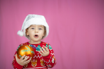 Fototapeta premium Cute child in a festive sweater and Santa hat holds a Christmas decoration on a pink background. The child is happy about the New Year and Christmas. Suitable for holiday projects.