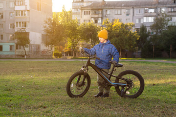 A 7-year-old boy standing beside his bicycle on a grassy stadium field with tall apartment buildings in the background. Dressed warmly in a jacket and hat, captured at sunset. Perfect for urban childh