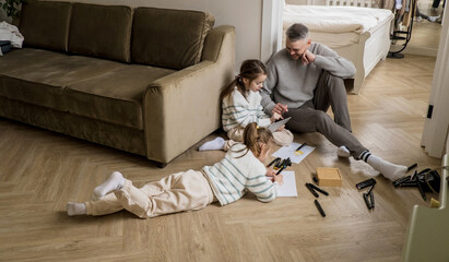 A father sits on the floor in his living room, watching as his two daughters engage in creative activities. One daughter is drawing with markers while the other is looking at a tablet with her father.