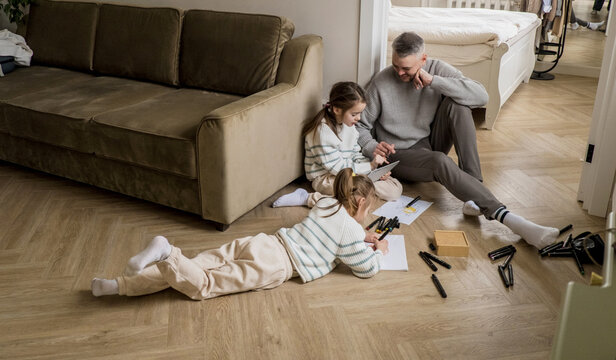 A father sits on the floor in his living room, watching as his two daughters engage in creative activities. One daughter is drawing with markers while the other is looking at a tablet with her father.