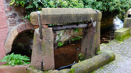 An ancient water wheel structure stands by a bubbling stream in a charming village, showcasing historical architecture surrounded by lush greenery and cobblestone pathways.