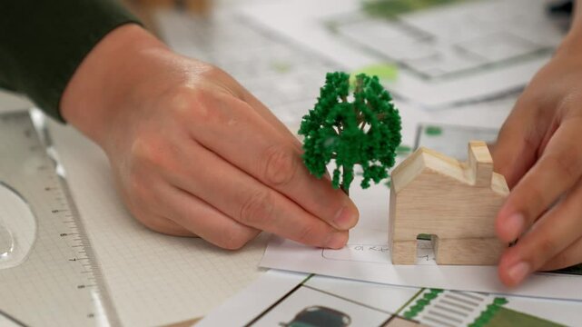 A video of architect using tree and home model to measure blueprint. Architect designing house construction on a table at studio, with architectural plan scattered around. Focus on hand. Delineation.