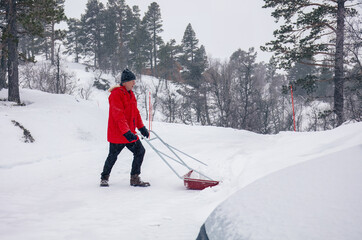 Man in red jacket pushing a snow pusher on a snow-covered road by mountain cabin ,snow removal,...