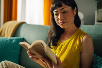 Asian woman deeply engrossed in reading a book on comfortable couch.