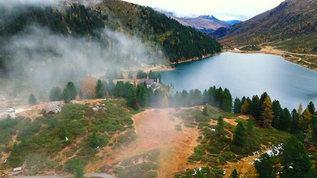 Aerial footage of mist hovering over Obersee Lake and a mountain lodge at Staller Sattel. The autumn landscape features colorful trees, calm water, and winding roads at the Austria-Italy border.
