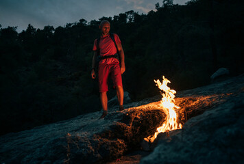 Obraz premium Hiker with backpack sits beside natural flames of Yanartaş, also known as eternal fire of Mount Chimaera, in Turkey. The night scene captures the unique fire phenomenon emerging from the mountain