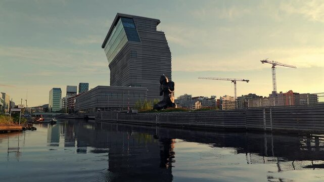 Sunset view of the Munch Museum and nearby sculpture in Oslo, with modern architecture and their reflections mirrored in the calm waterfront.