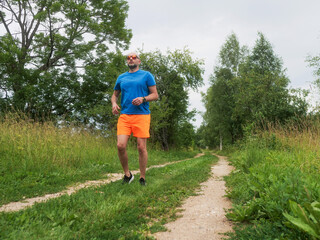 A man in a blue shirt and orange shorts walks down a dirt road. The man is wearing a hat and he is enjoying the outdoors. Male in his 40s with grey beard. Sport and fitness outdoor.