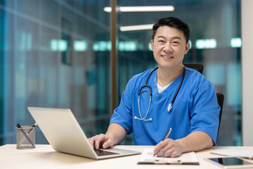 Asian doctor with stethoscope smiles confidently at desk, working on laptop in modern office. Portrays professional dedication and approachability in healthcare setting.