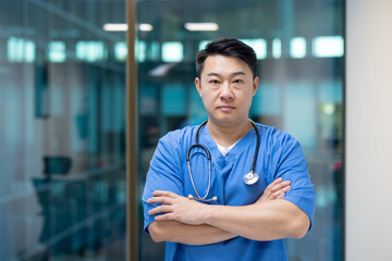 Fototapeta premium Asian doctor in blue scrubs with stethoscope, arms crossed, standing confidently in hospital. Represents professionalism, expertise, dedication in medical healthcare setting.