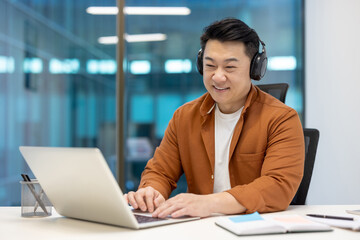Asian businessman with headphones working on laptop showcases focus, productivity in office. Casual...