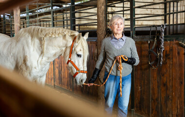 Elderly woman leading a white horse out of the stable