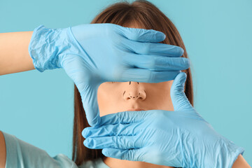 Fototapeta premium Beautiful young woman in gloves with marks on her nose against blue background. Plastic surgery concept