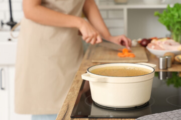 Cooking pot with soup on stove against housewife cutting carrot in kitchen, closeup