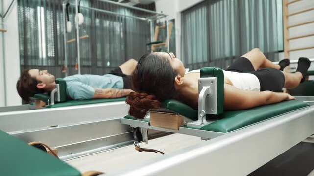 Couple practicing leg exercises on pilates reformer to strengthen core and improve flexibility at gym. Pilates partner doing pelvic bridge pose on reformer for improving lower back muscle. Habituate.