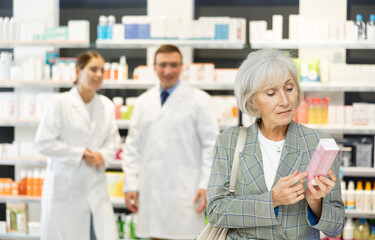 Near self-service window in pharmacy, senior female customer chooses cream for aging skin,...