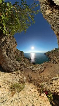 Vertical video. Wide Angle. A panoramic view from inside a natural rock formation overlooking the sunlit sea. The rugged cliffs and greenery frame the calm ocean, creating a beautiful and peaceful coa