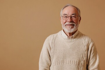 Fototapeta premium Elderly man in a casual sweater, smiling softly, standing against a solid beige background with plenty of space for text