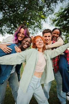 Vertical. Group of joyful young friends having fun outdoors, embracing and laughing together in a park. Energetic, carefree and multiracial people enjoying a lively and playful moment of friendship