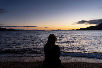 Fototapeta premium Una mujer sentada en la playa al atardecer, mirando el agua A woman sits on the beach at sunset, looking out at the water 