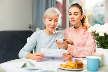 Mother and daughter sitting at table and calculating finances. Daughter using her smartphone for...